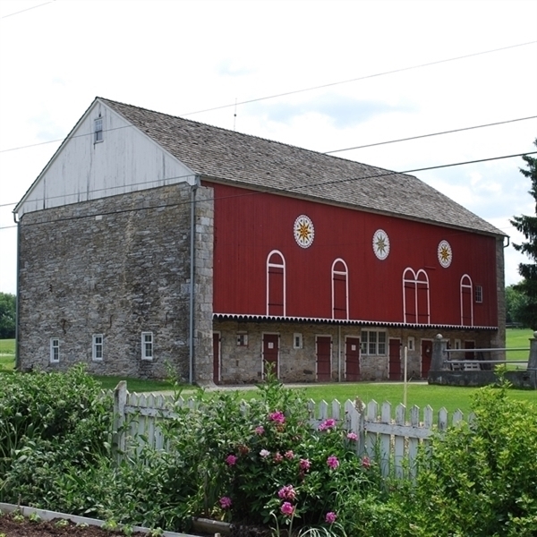 Image of the forebay side of the red and stone bank barn. The barn also has three barn stars painted along the side.