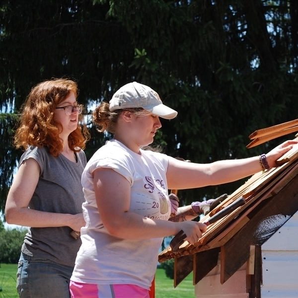 Two female identifying students working together to put on a wooden shingle roof on an outhouse