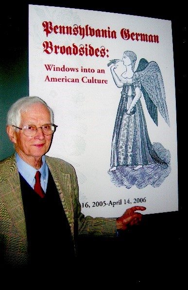 A white man with white hair and glasses stands in front of an enlarged cover of a book. He wears a blue shirt, a red tie, a dark vest, and a green suit jacket. He points to the enlarged cover of a book which features the title "Pennsylvania German Broadsides" in red German script.