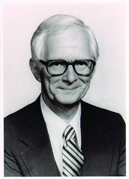 A black and white photo of a white man with white hair and dark rimmed glasses. He smiles at the camera and wears a light shirt, a striped tie, and a dark suit jacket.