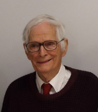 A white man with white hair and glasses smiles at the camera against a white background. He is wearing a white shirt, a red tie, and a dark sweater.
