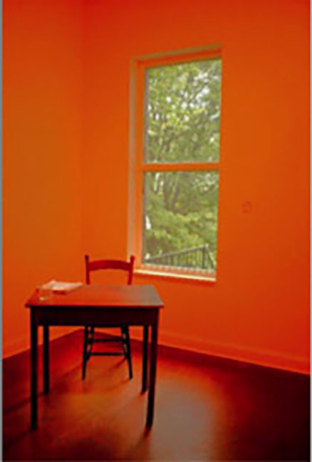 An image of a room, with orange walls, a chair and a table and a single window in the background