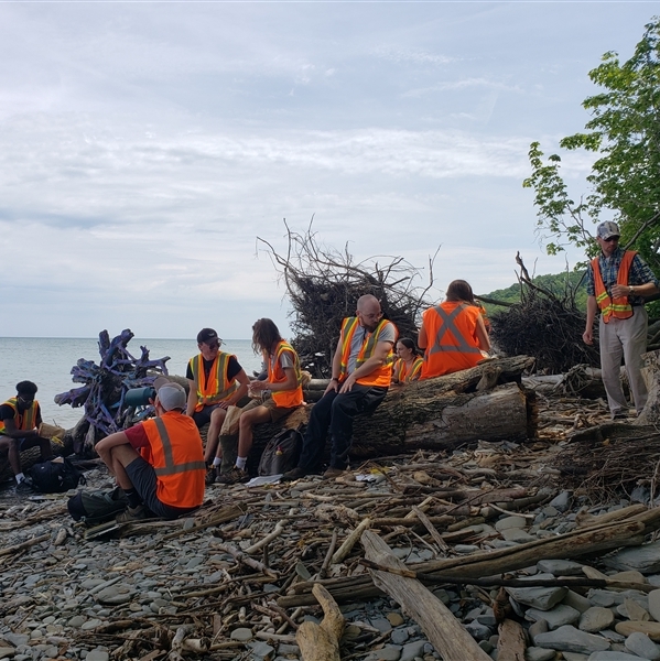 A group of students wearing neon orange vests, sitting on a fallen tree with the ocean in the background