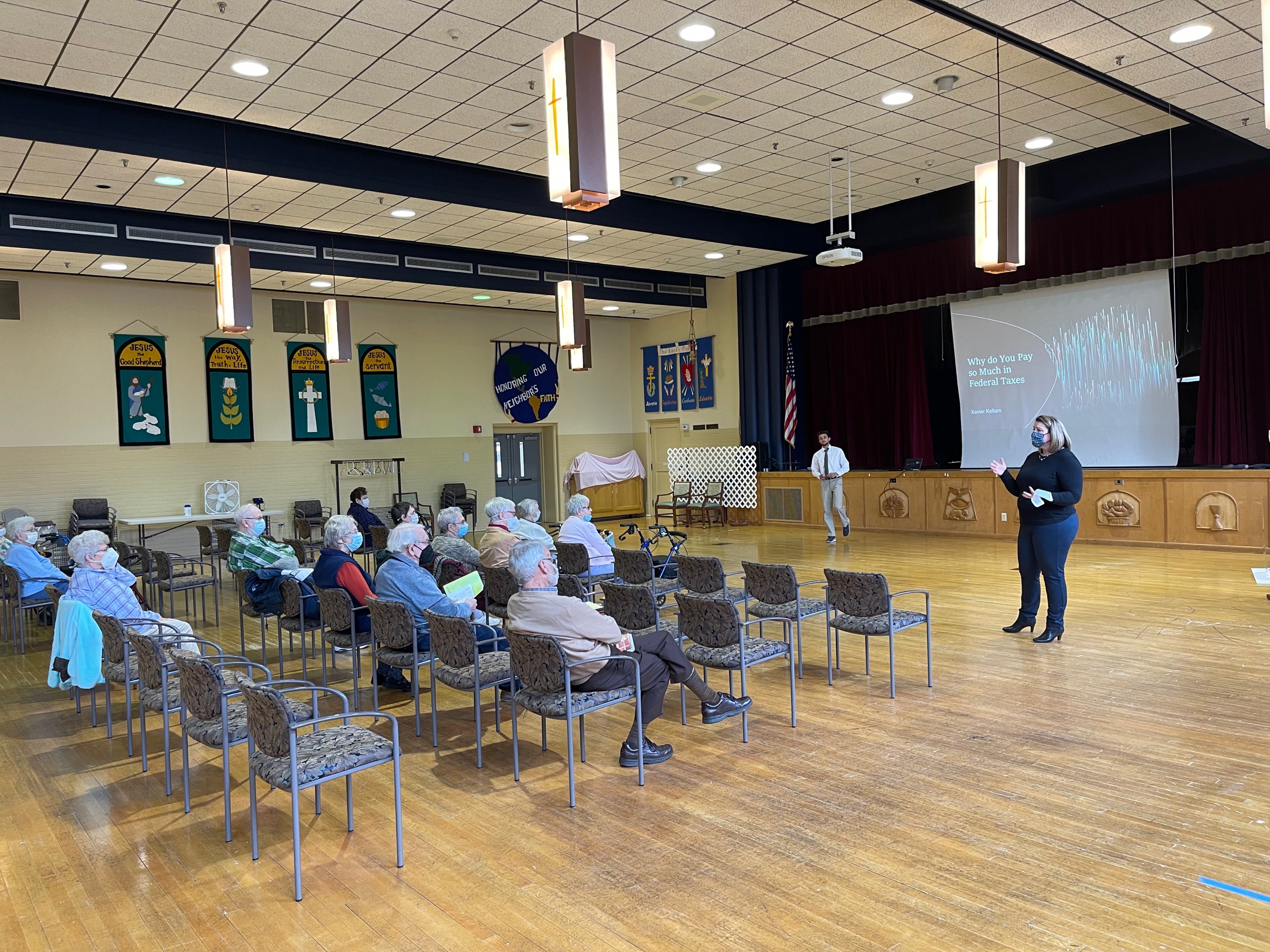 Topton community members seated facing right. Female student presenter in foreground facing left. Male student presenter in background. Projected screen with slide that says "Why do you pay so much in taxes?" behind student presenters.