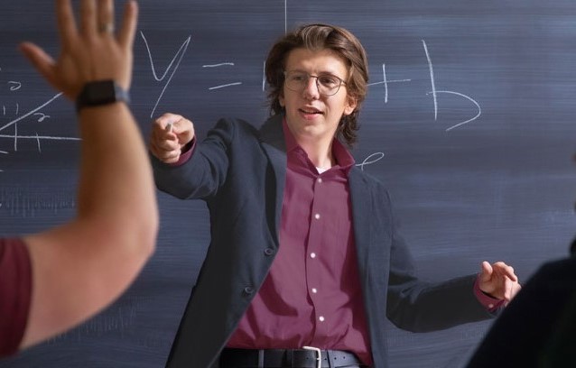 Student Michael Mister in a classroom, standing in front of a chalkboard with mathematic symbols, calling on a student who's hand is raised.
