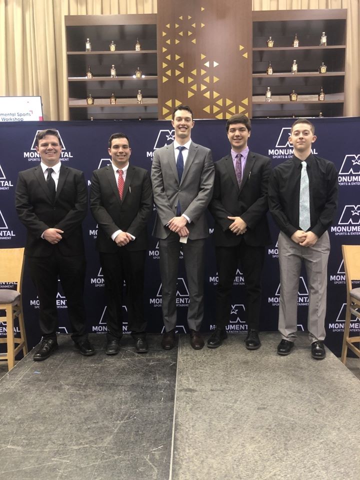five male students, dressed in suits, smiling and standing in a line at the Monumental Sports & Entertainment Sport Sales Workshop.