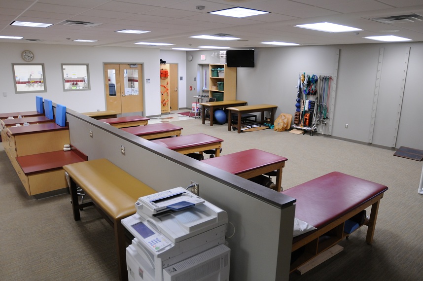 Wide shot of the training room with rows of medical tables and medical beds in the foreground, muscle training and therapy equipment and a flatscreen in the background 