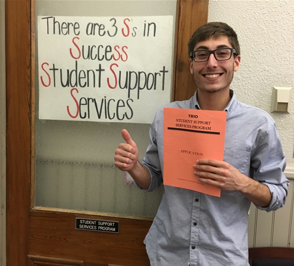 Male student hold TRIO application, in front of sign "There are three S's in Success.  Student Support Services.