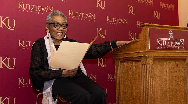 Crenshaw reading speech, seated beside podium.