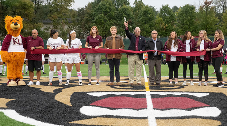 President Hawkinson cutting ceremonial ribbon in the middle of Keystone Field, surrounded by student athletes, faculty, and Avalanche, who are holding the ribbon for him to cut