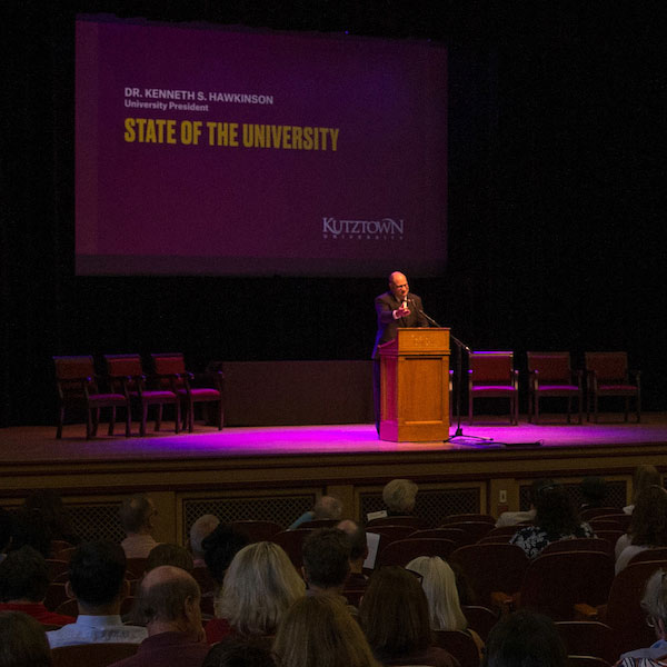 President Hawkinson at the podium, with the visual "State of the University" on the screen in the background, and the audience in the foreground.