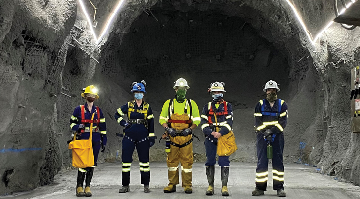 Alex and other miners stand in a large tunnel, wearing mining gear.