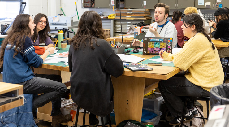 a group of student sitting around an art table designing on paper and laptops