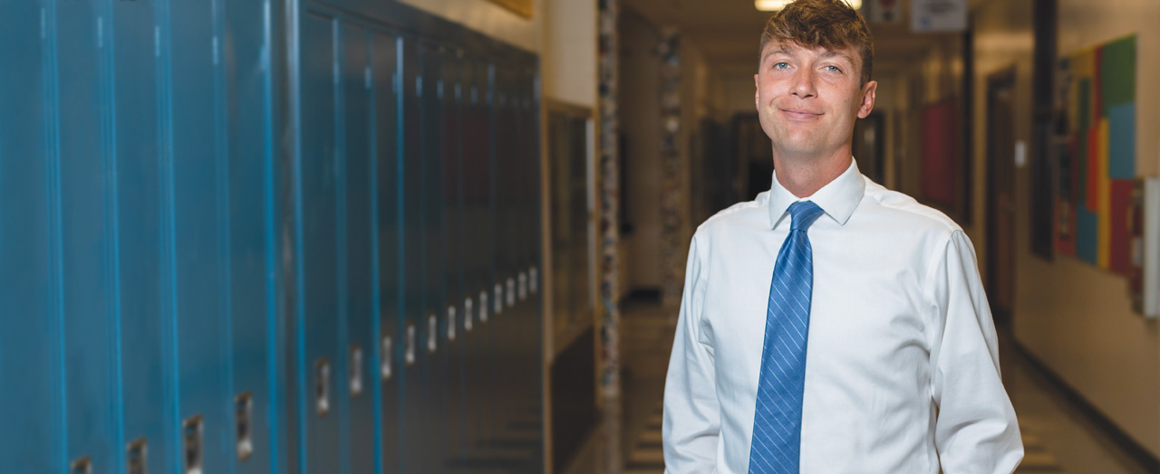 Joe Scoboria dressed professionally and smiling in an empty hallway next to a row of tall blue lockers 