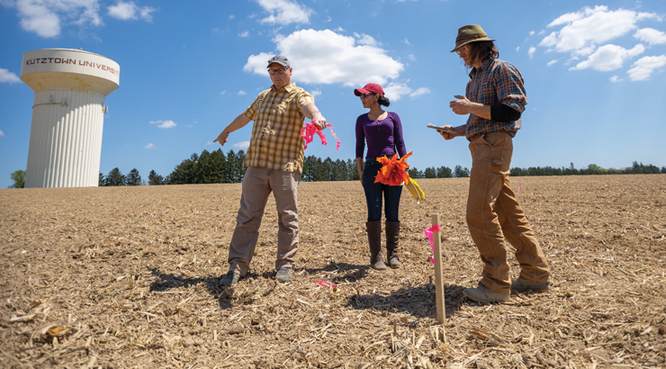 Three adults standing in a field in front of a marker taking notes and planning. The field is bare dirt against a blue sky and a water tower in the distance.