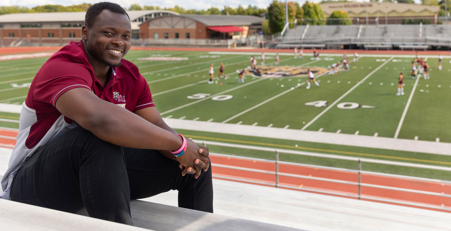 Sport Management major Rotuk smiling while seated on the stadium bleachers. In the background a team practices on the field.