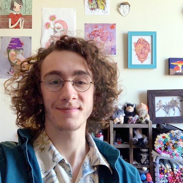 Image of Joey Strain with illustrations hanging on the wall behind him and art supplies on a shelf behind him.