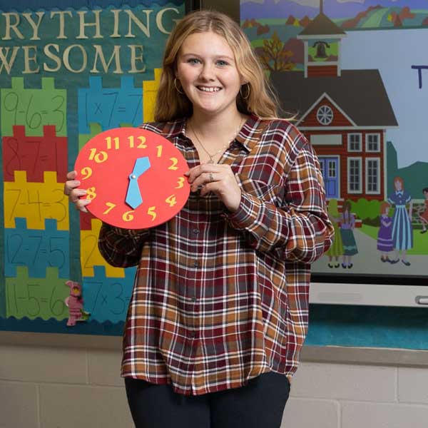 Female elementary education student standing in front of a digital school board holding up a clock
