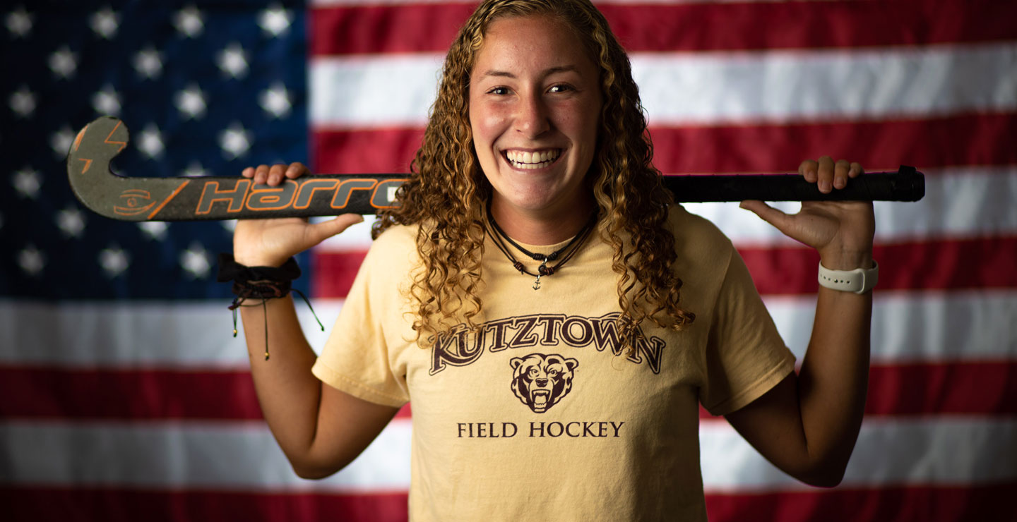 Erika Barbera pictured in front of an American flag, while holding a field hockey stick behind her, wearhing a Kutztown Field Hockey shirt.