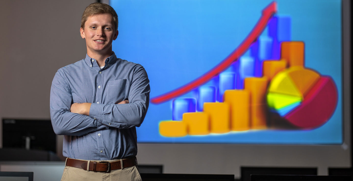 Male student looks into the camera, standing in front of a colorful projection on the wall with an arrow moving upward, blue and yellow chart and a multi-colored pie chart.