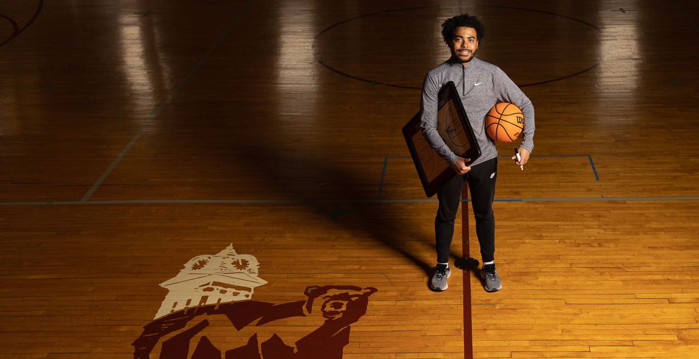 Male sport management student holding basketball playboard, marker and basketball on a wooden court, the old Golden Bear logo can be seen in the foreground.