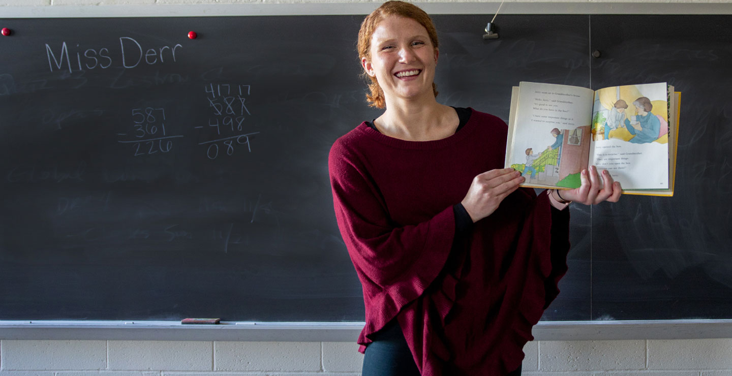 Female student in front of a classroom chalboard smiling and holding a children's picture book. Her name, Miss Derr, and a math problem are visible on the chalkboard.