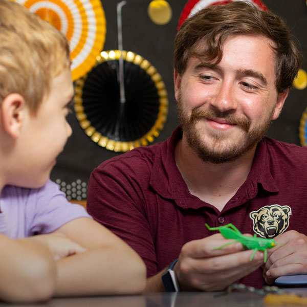 Male education major holding a green dinosaur, looks and smiles at a student he is teaching in a classroom setting. backgrround are math formulas and questions.