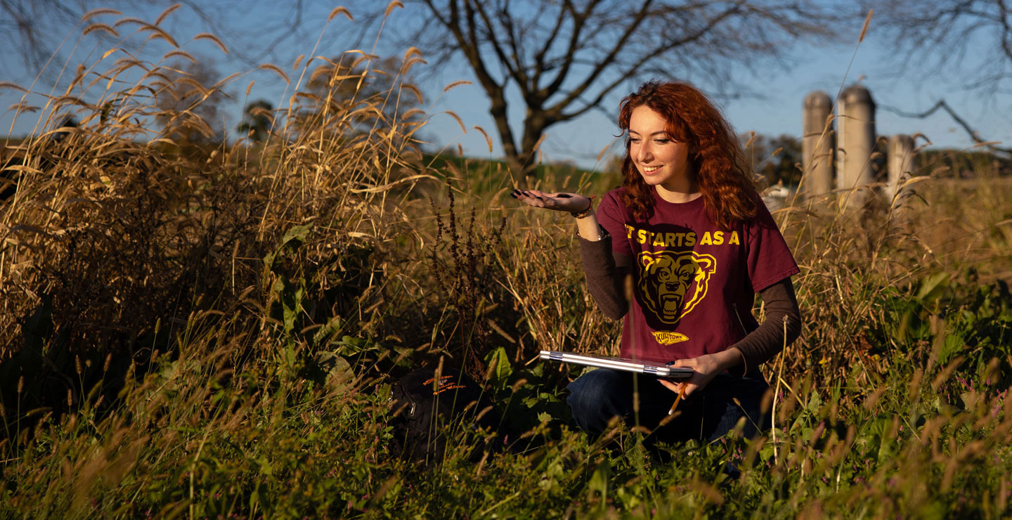 Female student, wearing a maroon KU shirt that reads "It starts as a" with a logo of the Golden Bear underneath.  She is holding a caterpillar in one hand and a laptop computer in the other.  In the background are silos of a farm, and she is surrnounded by plants and tall grass.