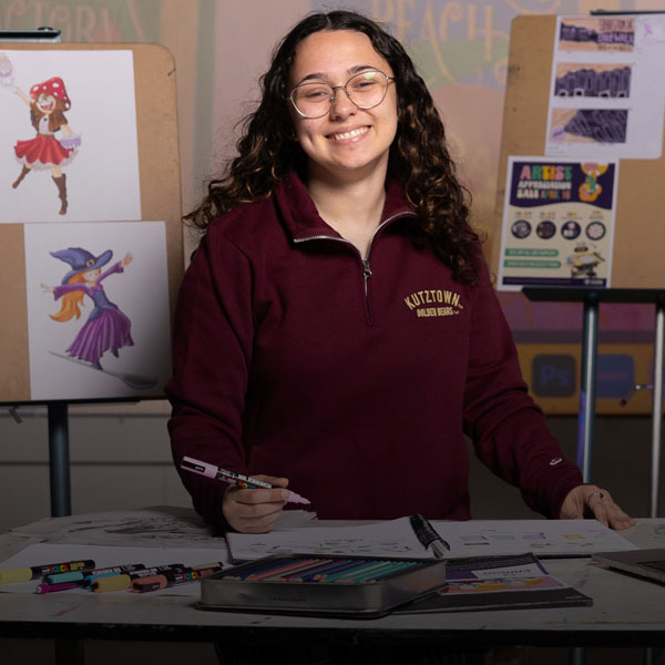 Female communication design major holding a colorful marker, while an array of markers is spread out in front of her.  Behind and in front of her are a series on artistic renderings and artwork on display.