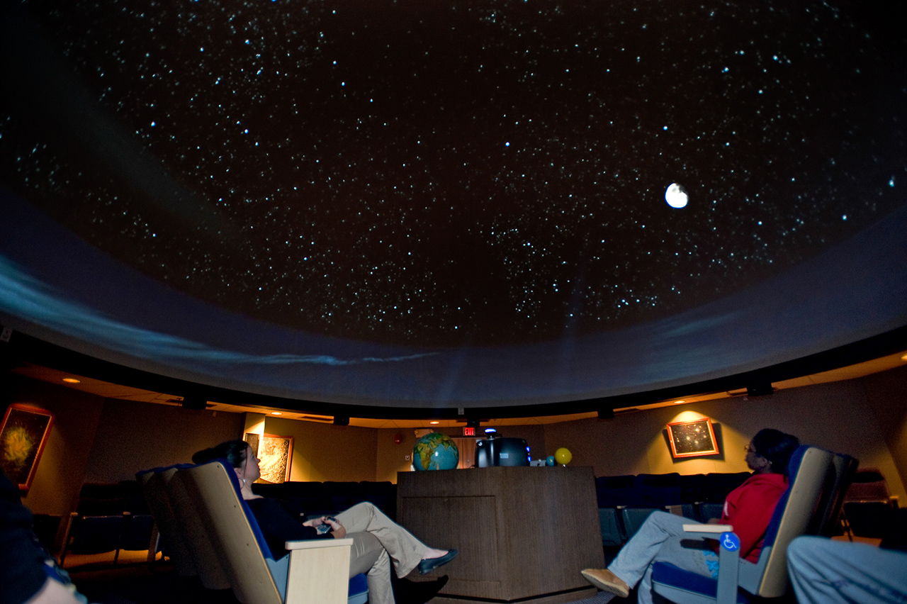 Planetarium in the grim science building with students sitting in reclining chairs, gazing at star show