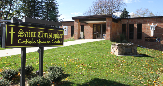 Distant shot of St. Christopher Catholic Newman center front entrance, with the welcome sign in the foreground 
