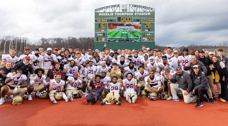 Football team poses in front of scoreboard showing the final score of KU's 28-16 win over Slippery Rock in Super Region 1 Championship