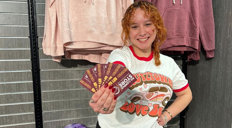 Female student wearing KU gear, standing in front of KU merch, holding Campus Store gift cards