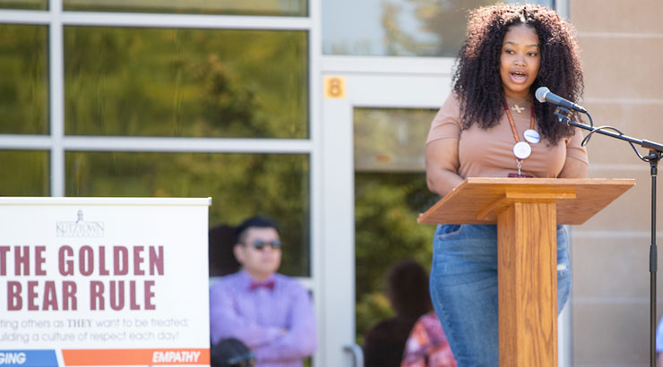 Female student at a podium, speaking into a microphone at a recent Unity Day Rally.  A sign nearby reads "The Golden Bear Rule"