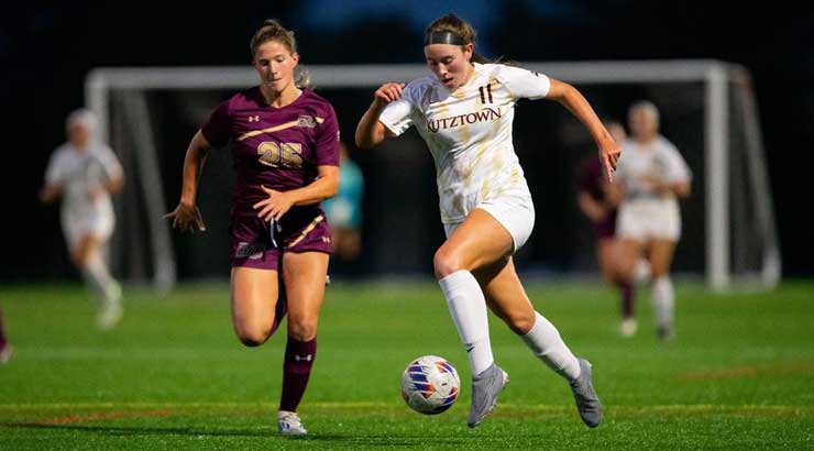 KU women's soccer player in a white uniform ready to kick the ball as she avoids a defender in a maroon uniform.