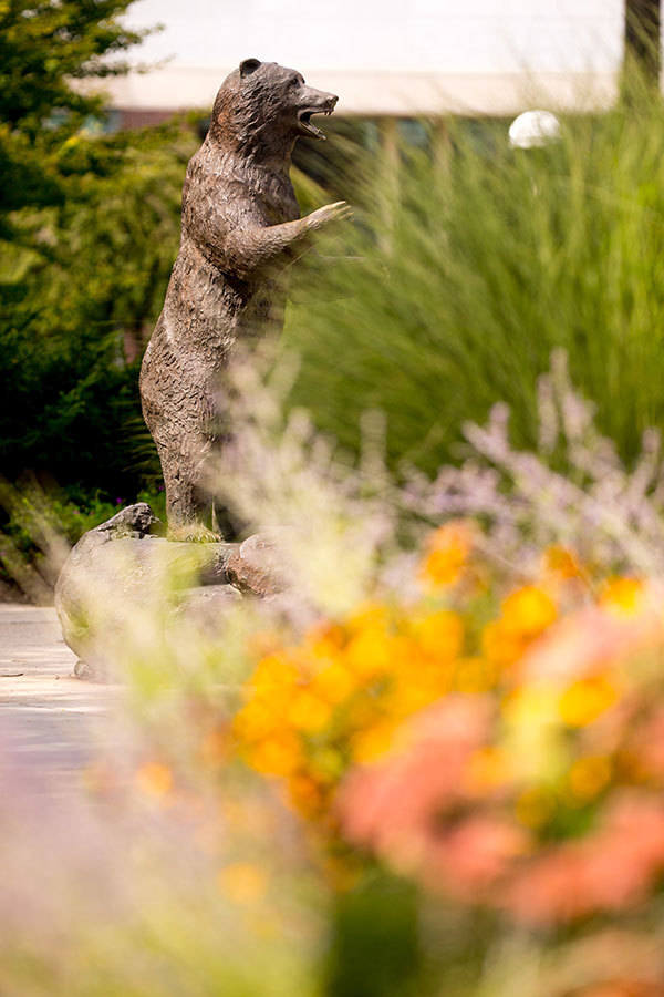 Metal statue of an upright bear surrounded by lush flowering gardens and trees