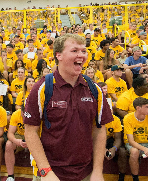 A while male student laughing and very enthusiastic in front of bleachers full of students all wearing spirit gold KU shirts at convocation celebrations