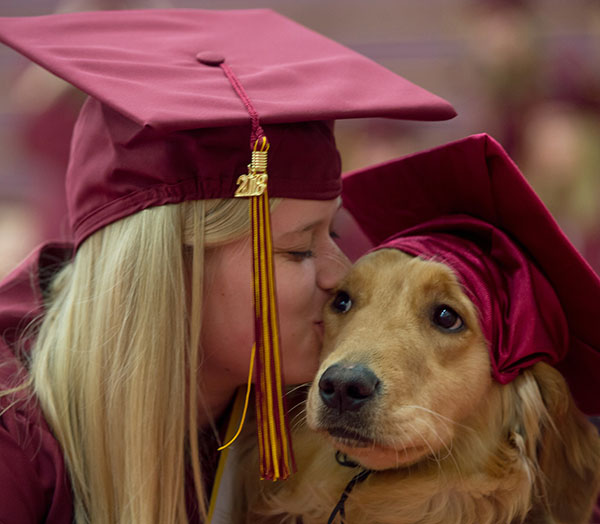 A female student with long blonde hair wearing a graduation cap leaning over to kiss a golden retriever also wearing a graduation cap