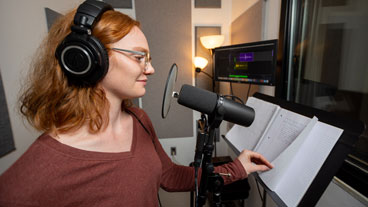 Female student wearing headphones stands in front of a microphone looking at a script.