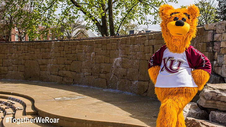 Avalanche the golden bear standing with his hands on his hips in front of the fountain wall on the alumni plaza