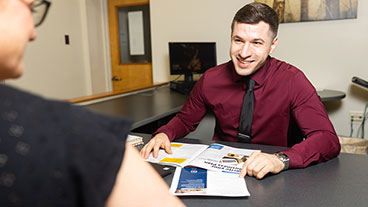 Male student from Master of Business Administration program sitting at desk, explaining MBA degree to another student.