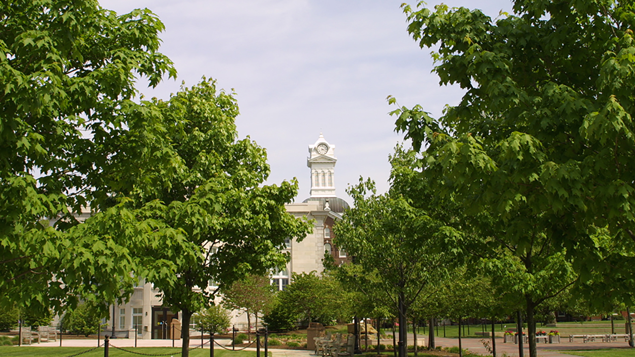 Old Main clock tower at a distance, the entrance of Old Main obscured by the leafy branches of nearby trees