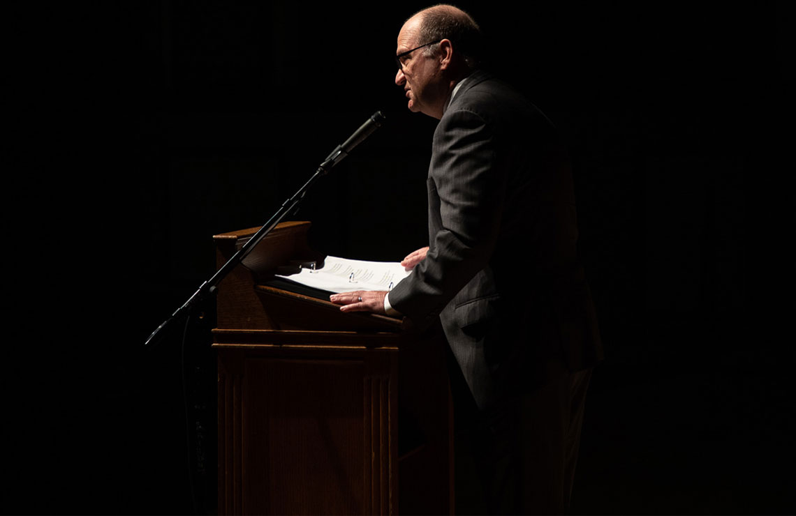 President Hawkinson standing at a podium on a dark stage with a spotlight lighting just him speaking into a microphone