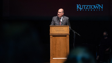President Hawkinson speaks from behind podium, with "Kutztown University" visibile on the screen behind.