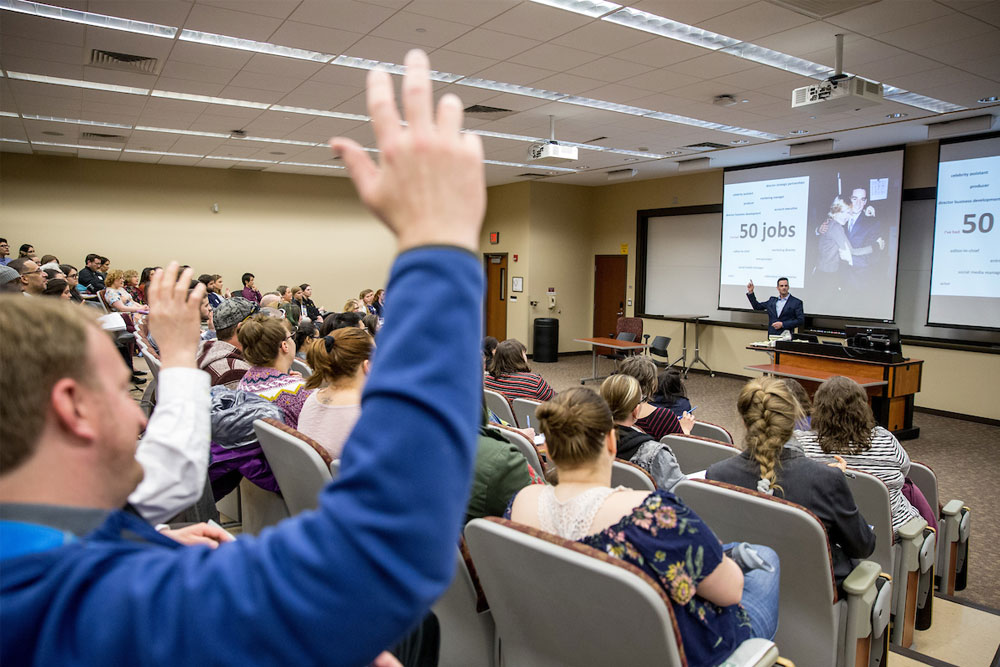 Students in classroom with hands raised, professor at front of the room in front of screen with words and photos.