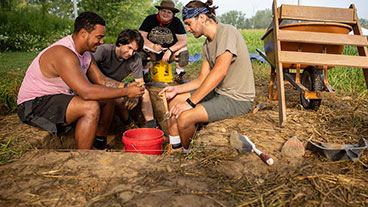 A group of three male and one female students of diverse ethnic backgounds at a archaeological dig site.