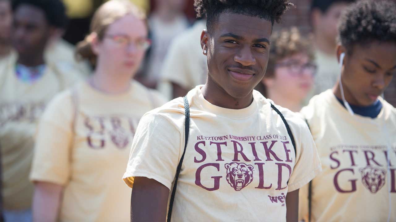 A small crowd of newly appointed golden bears, all wearing a shirt stating strike gold. An african american gentelman is smiling at the camera as the main focus of this image.